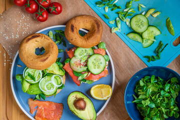 Homemade bagel sandwiches. Gluten free toasted bagels with smoked salmon and cream cheese and with avocado, cucumber, lettuce, and onions. Healthy breakfast on wooden board, fun vibrant colors