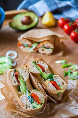 Stuffed toasted bagel sandwich cut into half with avocado, mayo, cucumber, lettuce and tomatoes. Salmon bagel in the background, wrapped with paper. Gluten free healthy breakfast, vibrant fun colors 