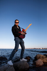 A guy with a red electric guitar in a leather jacket on the background of the lake stands on the stones and plays rock. Rock guitarist in sunglasses plays against the background of water and sky stand