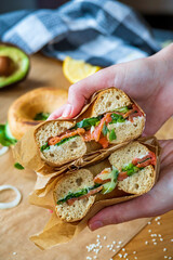 Hands holding stuffed toasted bagel sandwich cut into half with smoked salmon, cream cheese, cucumber lettuce. Avocado bagel in the background. Gluten free healthy breakfast, vibrant fun colors