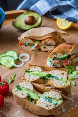 Stuffed toasted bagel sandwich cut into half with avocado, mayo, cucumber, lettuce and onions. Salmon bagel in the background, wrapped with paper. Gluten free healthy breakfast, vibrant fun colors