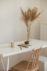 Women's home office workspace. Freelance work, business workplace. Light aesthetic hygge space with wooden chair, table, reed pampas grass bouquet, mug, notebook. Modern interior design.