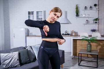 Fototapeta na wymiar Young beautiful woman in leggings and a top shows a fold on the belly. Healthy lifestyle. The woman goes in for sports at home. Fighting excess weight at home