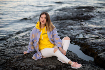 A beautiful girl in a coat and white pants with a hole in her knee sits on a rocky shore and thinks about something. Evening portrait of a stylish young woman outdoors.