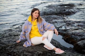 A beautiful girl sits on a rocky shore and thinks about something. Evening portrait of a beautiful young woman in nature.