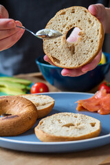 Making homemade bagel sandwich. Gluten free toasted bagel with hands spreading cream cheese and adding salmon, lettuce, cherry tomatoes, avocado and cucumber. Healthy breakfast with fun vibrant colors