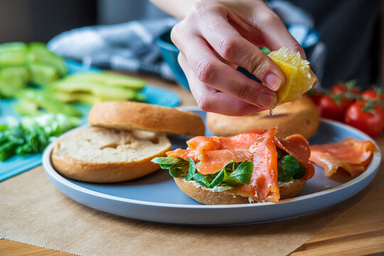 Bagel sandwiches in the making. Gluten free toasted bagel with smoked salmon and cream cheese. Hand squeezes lemon on top with avocado and tomatoes in background. Healthy breakfast, fun vibrant colors - Powered by Adobe