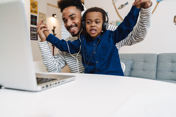 Happy african american father and son in headphones using laptop at home