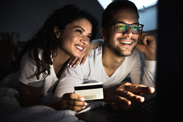 Couple lying on bed shopping online via laptop at night.