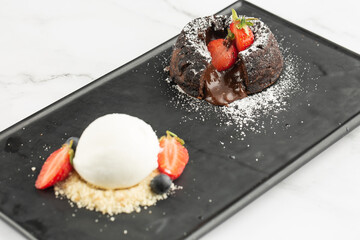 Chocolate fondant with vanilla ice-cream and berries. Hot chocolate pudding cake served in a black slate rectangular plate. Isolated close-up on a white marble background. Dessert menu concept.