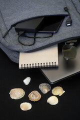 A small gray textile backpack with a laptop or ultrabook, a smartphone, sunglasses and an external hard drive lies on a black surface next to seashells. Modern traveler's gadgets