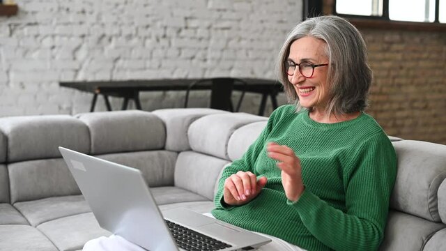 Cheerful senior woman has a video conference on the laptop sitting on the couch at home, an elderly lady wearing eyeglasses staying in touch with her friends or family via video connection, video call
