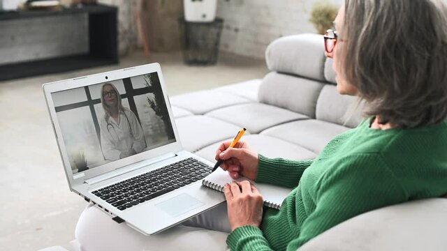 Side view the senior woman has a video consultation on the laptop, online video appointment with a doctor, an elderly female patient get online recommendation from a kaiser therapist, taking a notes