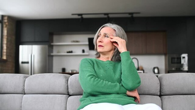 Frustrated senior woman feels headache, migraine, a mature retired lady sits on the couch at home and holds forehead, a worried grandmother has memory losses, trying to remember smth