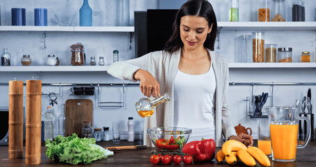 Brunette woman pouring oil in salad near fresh ingredients in kitchen