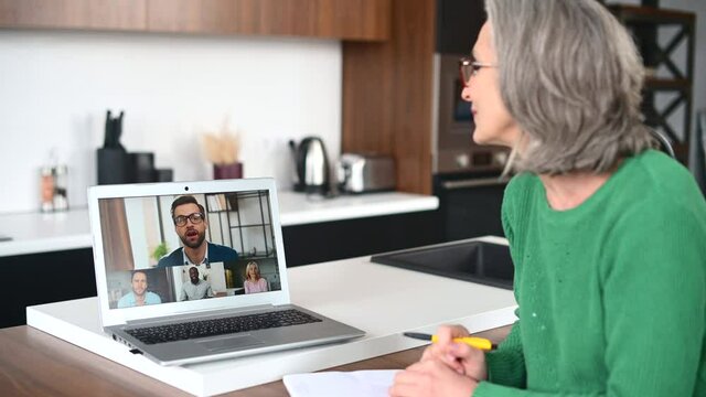 Back view of a senior female tutor, coworker wearing a headset is talking via video connection with a group of diverse people on a laptop screen. Mature woman is using computer app for teaching online