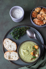 Broccoli cream soup decorated with thyme, cream, roasted cauliflower and fresh bread on green concrete background. Healthy vegetarian food concept. Top view.