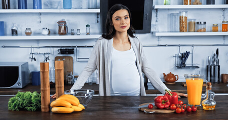 Pregnant woman looking at camera near fresh vegetables and orange juice