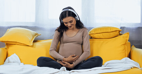 Smiling pregnant woman in headphones embracing belly in living room