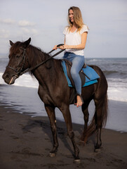 Beautiful woman riding horse on the beach. Outdoor activities. Caucasian woman wearing jeans and white T-shirt. Traveling concept. Cloudy sky. Bali, Indonesia