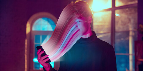 Cinematic portrait of stylish man in neon lighted interior using a smartphone. The face is smeared, sucked, absorbed into the phone. Concept of social network dependency, phone addiction. - obrazy, fototapety, plakaty