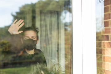 A young man with a face mask on looking and waving out the window during his self-isolation at...