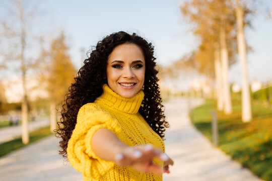 Girl with curly dark hair European smiling reaches out her hand to the camera standing on the alley in the park. High quality photo