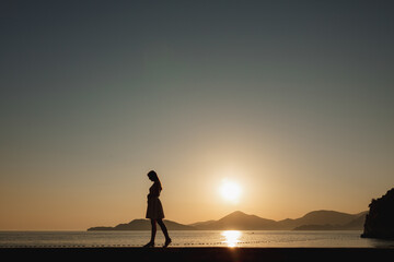 A pregnant woman walks on the seashore at sunset and gently strokes her belly, behind her are mountains and the setting sun 