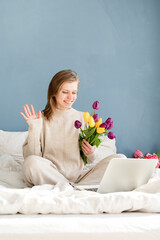 Happy woman sitting on the bed wearing pajamas holding tulip flowers bouquet working on laptop