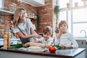 Cheerful and joyful boys and their young mother cook a pizza and learn to cook in kitchen in daytime.