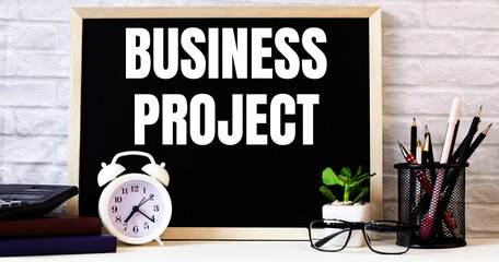 The word BUSINESS PROJECT is written on the chalkboard next to the white alarm clock, glasses, potted plant, and pencils in a stand.
