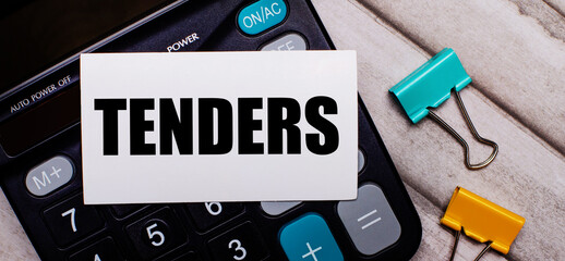 Calculator, multi-colored paper clips and a white card with the inscription TENDERS on a light...