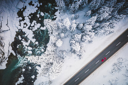 Car Traveling On The Snowy Mountain Road On Side Of Frozen River And Woods, Aerial View, Switzerland, Europe