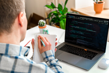 Over his shoulder, a close-up view of a man writing in a notebook, a programmer using a laptop...