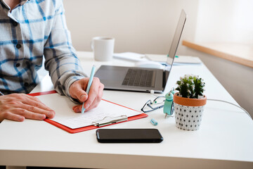  A young man in a shirt in a bright office works at a laptop and makes notes in a notebook, the...