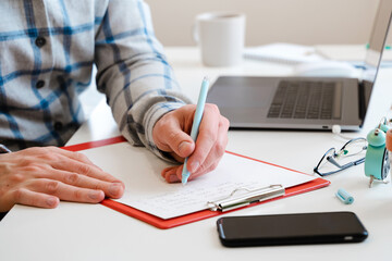  A young man in a shirt in a bright office works at a laptop and makes notes in a notebook, the mental activity of a project manager or an application developer