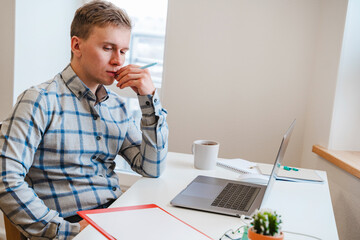 A young man in a shirt in a bright office works at a laptop and makes notes in a notebook, the mental activity of a project manager or an application developer