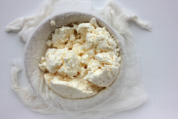 Close-up of freshly made white soft cottage cheese in bowl on white table background. Homemade ricotta in cheesecloth. Top view
