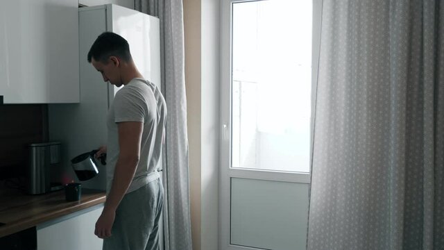 Handsome young man pours coffee from coffee maker and walks up to window in kitchen.