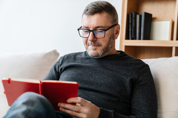 Pleased adult unshaven man writing down notes while sitting on couch