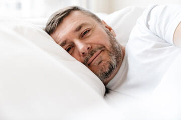 Peaceful adult unshaven man looking at camera while lying in bed