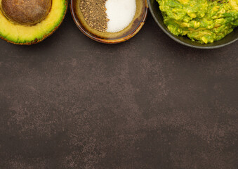 Top view of fresh guacamole on a dish placed on a dark gray stone background with ingredients for homemade guacamole: avocado, salt, and pepper. Concept of traditional Mexican preparation