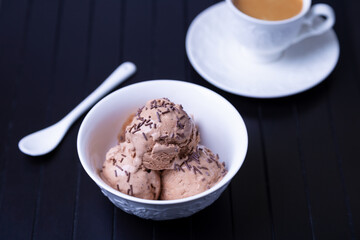 Chocolate ice cream with chocolate sprinkle in a white bowl on a black background. Three balls of homemade ice cream and a cup of coffee. Close-up.