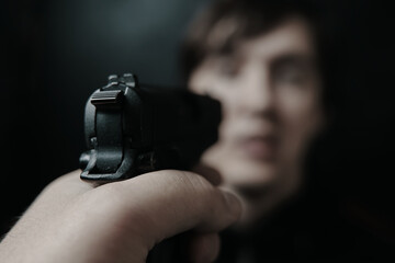 First person view of gun aimed at young man on black background. Close up of criminal with firearm. Murderer or armed thief. Pistol in man's hand. POV of aiming at human target.