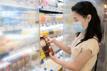 Asia woman wearing medical face mask and pick up milk from the refrigerator shelf. Reading nutritional value from the milk bottle. Shopping at supermarket in new normal style during coronavirus