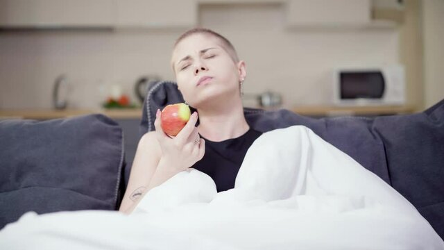 Portrait of young Caucasian hairless woman eating healthful apple sitting on couch at home. Ill female patient recovering after chemotherapy. Hope and health care concept.