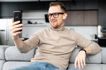 Young bearded hipster guy in glasses and casual clothes wearing wireless earphones sitting on a...