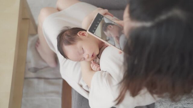 top view, asian woman parent holding baby infant using mobile smart phone taking photo of a kid in living room at home. kid sleeping in mom arm. natural authentic.