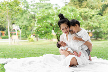 Asian young beautiful mother holding her newborn is sleeping and feel with love and touching gently then sitting on green grass in the park