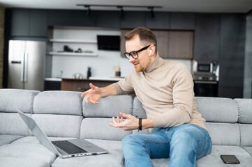 Fototapeta premium Handsome fit young bearded hipster man in glasses and wireless earphones sitting on the couch, talking with emotions and gesturing, video-calling via laptop online, male freelancer talking on webcam